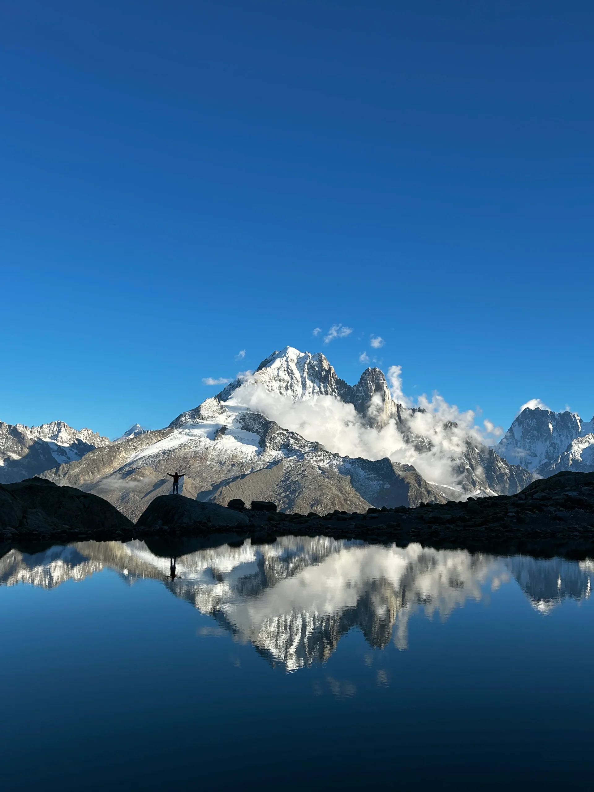 Panoramic view of snow-covered mountains reflected perfectly in a calm alpine lake under a clear blue sky at Refuge du Lac Blanc.