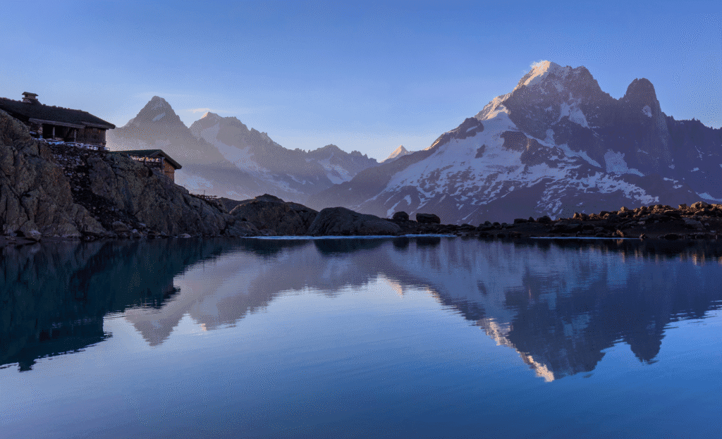 Morning view of Refuge du Lac Blanc and the snow-covered peaks of the Mont Blanc range reflected in the still alpine lake.