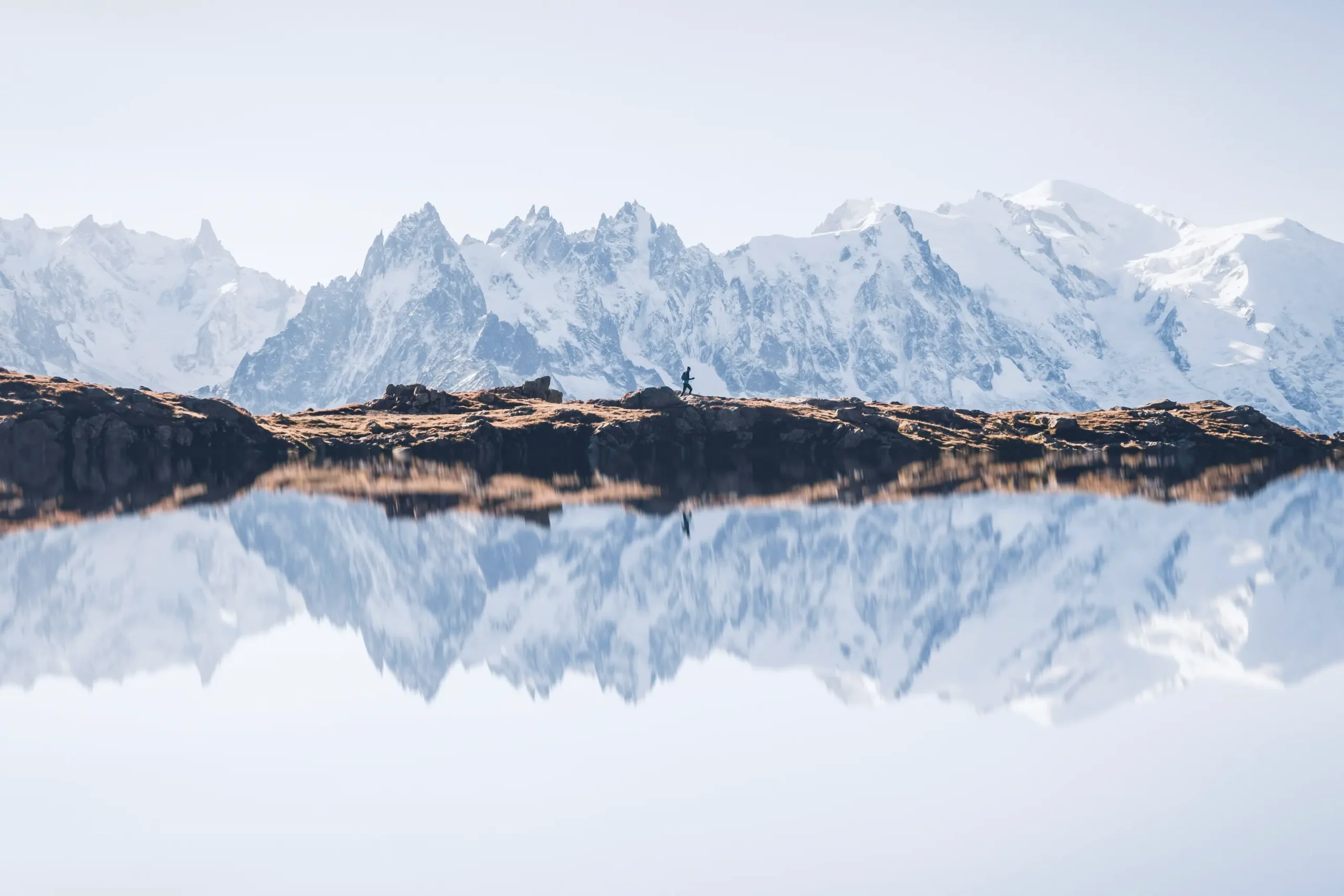 Randonneur au bord du Lac Blanc face aux aiguilles enneigées du massif du Mont-Blanc, reflet des montagnes dans l’eau calme près du Refuge du Lac Blanc à Chamonix